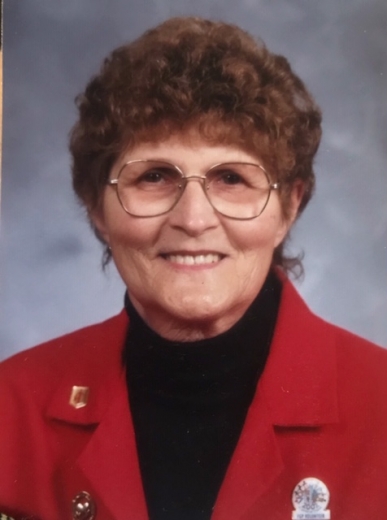 A professional portrait of Ruth. She smiles, wearing a red jackets and wire-framed hexagonal glasses.