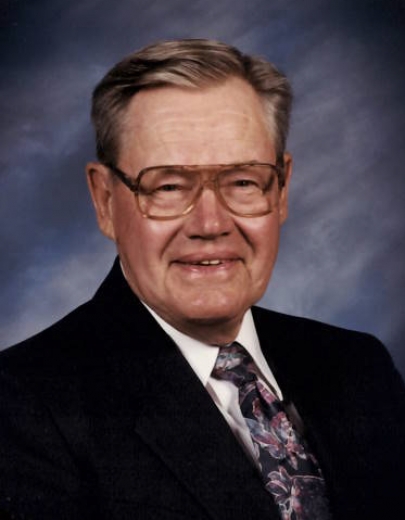 Andre Torkelson smiles in front of a blue background. He is wearing a suit and tie and wire-rimmed glasses.