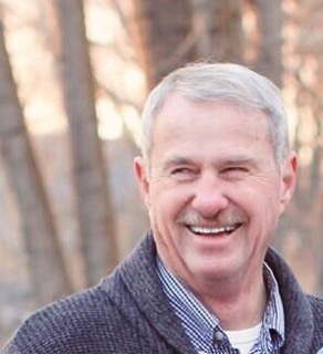 Pat laughs. Behind him are bare trees and he wears a blue cardigan.