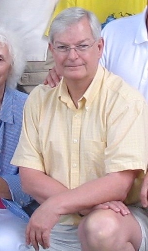 A photo of Ray kneeling, cropped from a group photo. He wears glasses and a pastel yellow polo shirt.