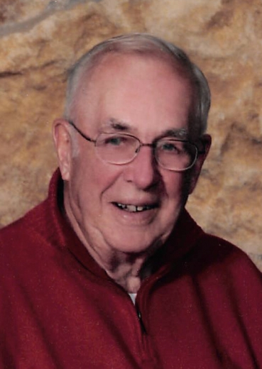 LeRoy smiles, wearing a red sweater in front of a mottled brown background.