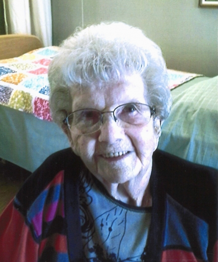 Lillian Halverson smiles, wearing a color-block sweater in black, blue, red, and magenta, and a gray blouse with flower on it. Behind her is a care center room with a quilt on the bed.