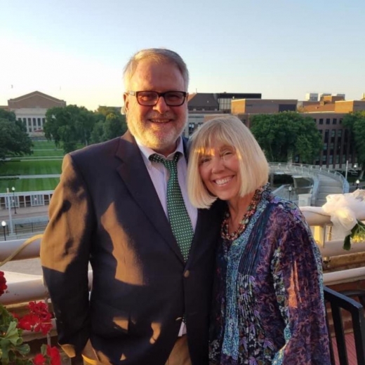 Steve and Diane stand arm in arm. They are dressed up, and behind them are trees and buildings.